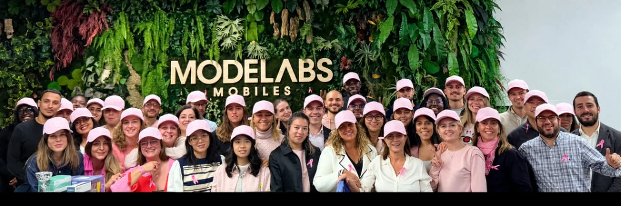 Photo de groupe des collaborateurs Modelabs devant le mur végétal du siège, coiffés de casquettes roses